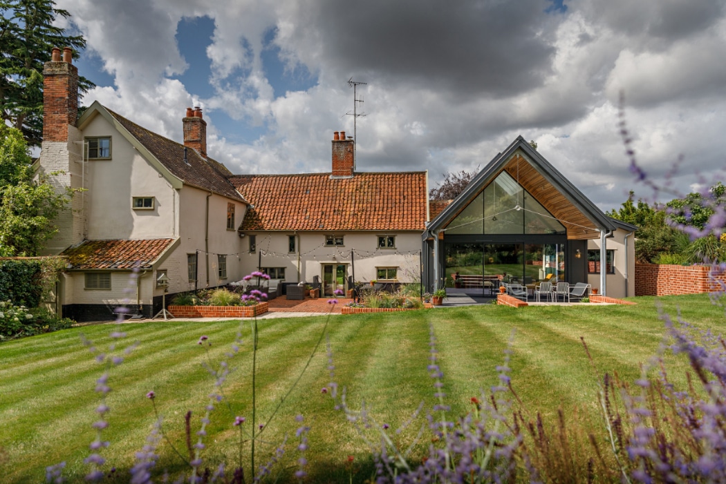 A traditional house with tiled roofs and brick chimneys features a modern glass extension, set on a well-kept lawn with cloudy skies overhead.