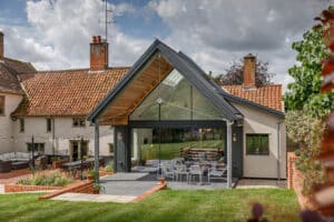 Modern glass extension with sloped roof attached to a traditional house, featuring outdoor seating and a large lawn in the background.