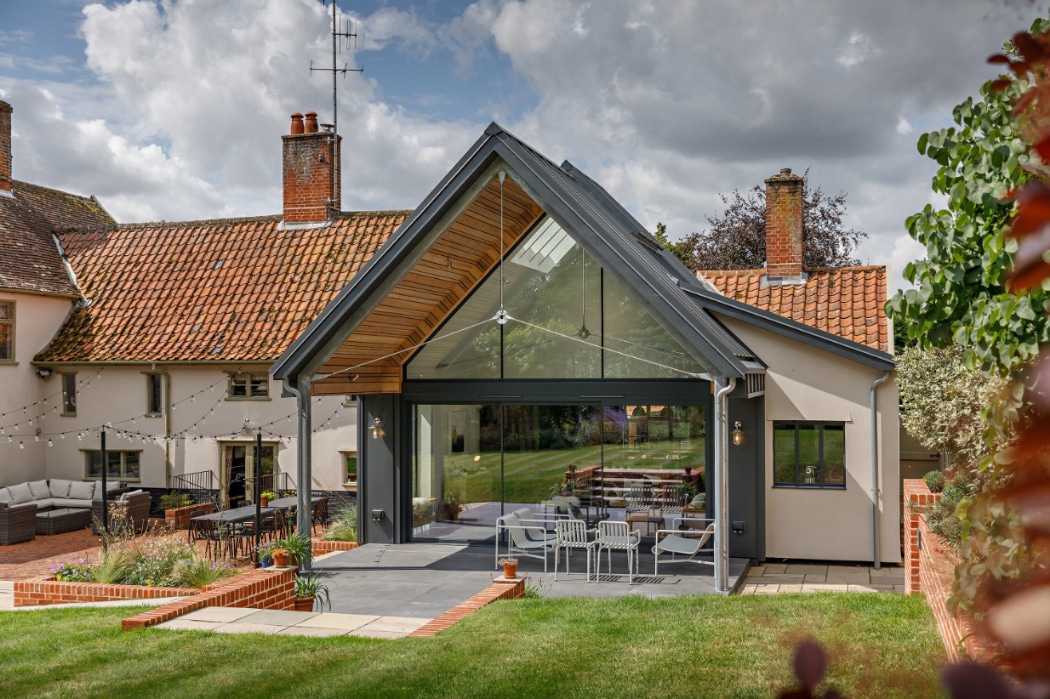 Modern glass extension with sloped roof attached to a traditional house, featuring outdoor seating and a large lawn in the background.