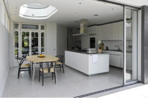 Modern kitchen with white cabinets, central island, and dining table with four chairs; glass doors and skylight provide natural light.