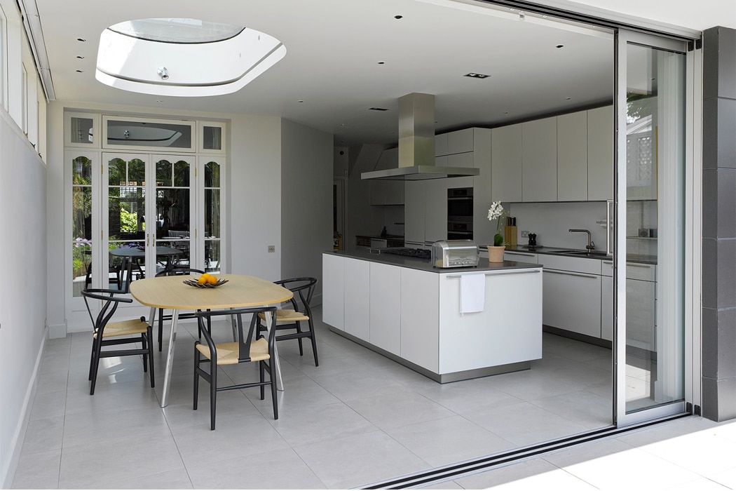 Modern kitchen with white cabinets, central island, and dining table with four chairs; glass doors and skylight provide natural light.