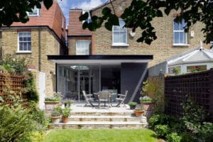 A modern glass extension opens onto a patio with outdoor dining furniture, surrounded by plants and wooden fencing, at the back of a brick house.