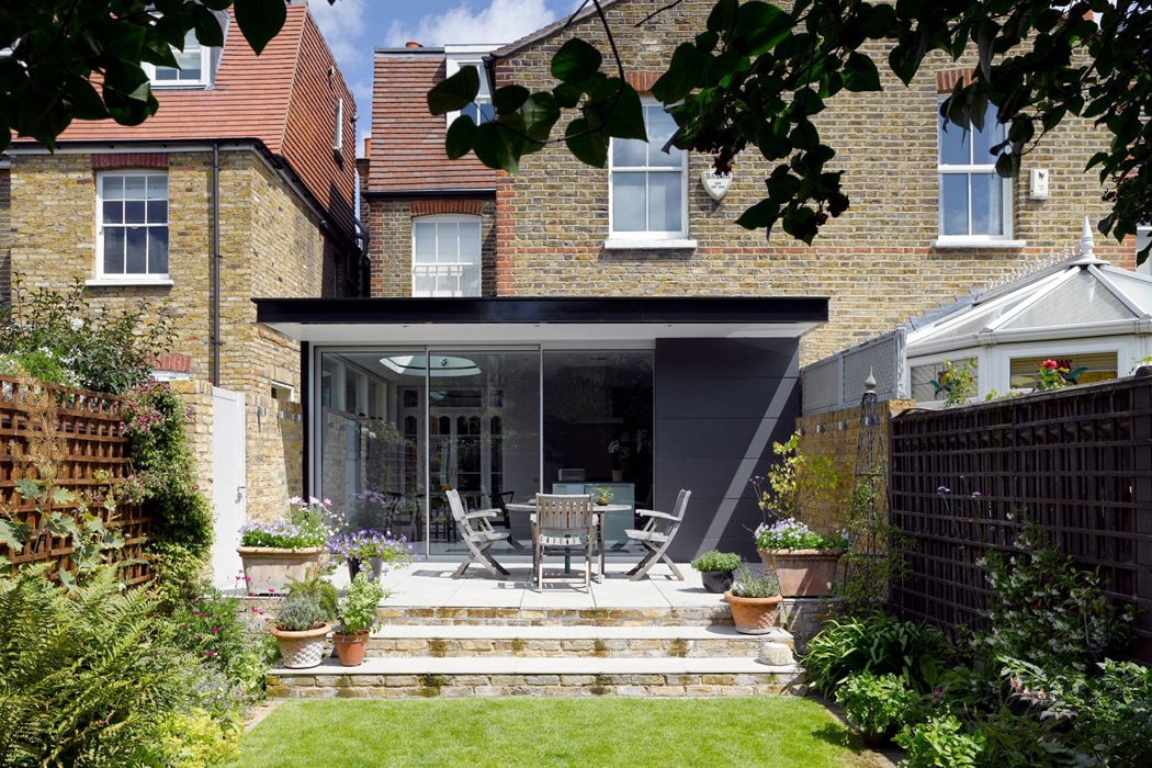 A modern glass extension opens onto a patio with outdoor dining furniture, surrounded by plants and wooden fencing, at the back of a brick house.