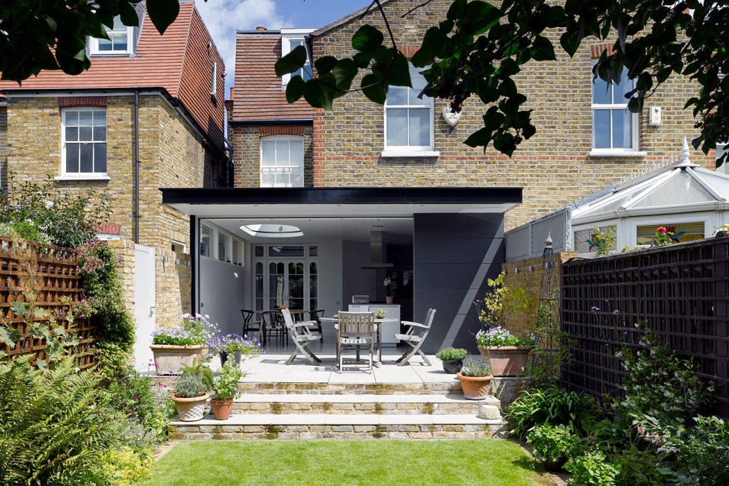 A modern garden patio with outdoor seating and dining area extends from the back of a brick house, surrounded by plants and wooden fencing.