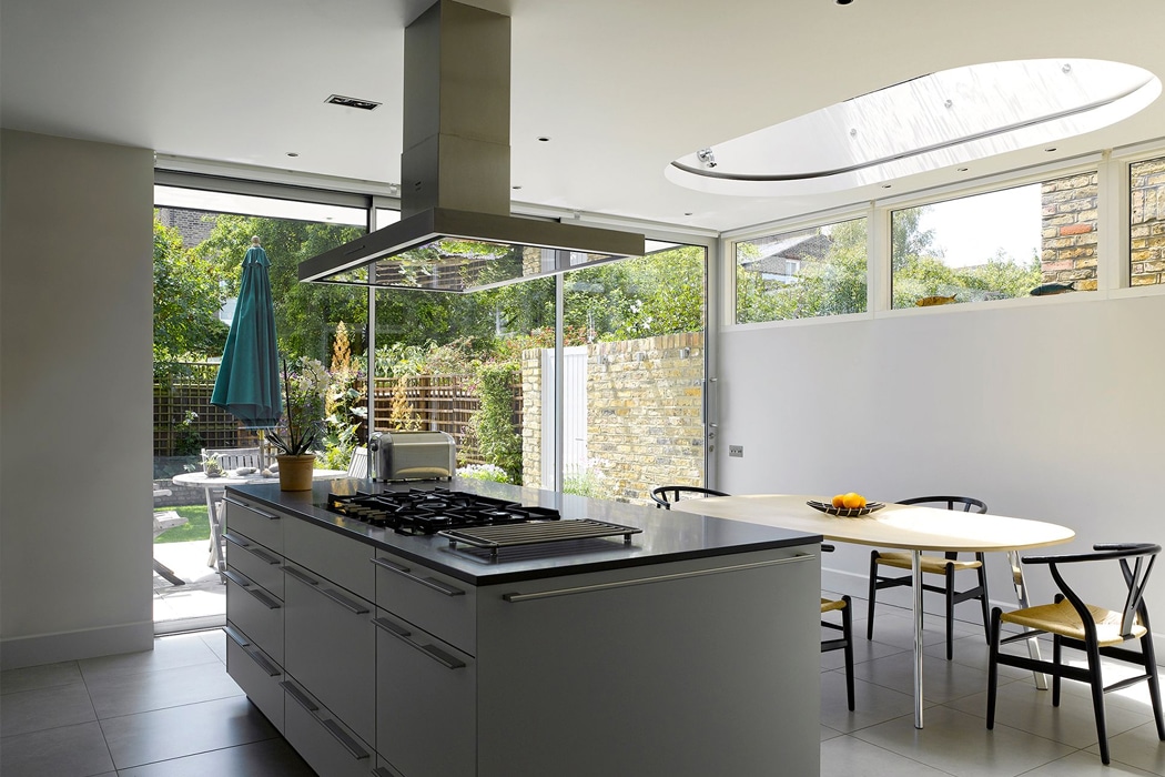 Modern kitchen with a central island, built-in stove, dining table with chairs, large windows, and a view of a green backyard. Natural light fills the space.