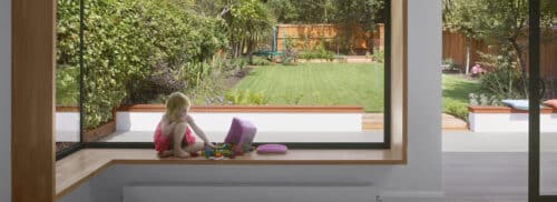 A girl sitting by a frameless glass-to-glass corner window in a modern extension, offering uninterrupted panoramic views.