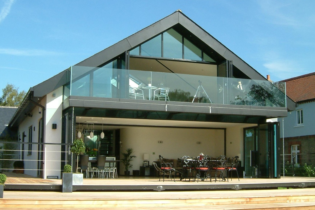 A modern two-story house with large glass windows, a spacious balcony with glass railing, and outdoor dining furniture on a wooden deck.