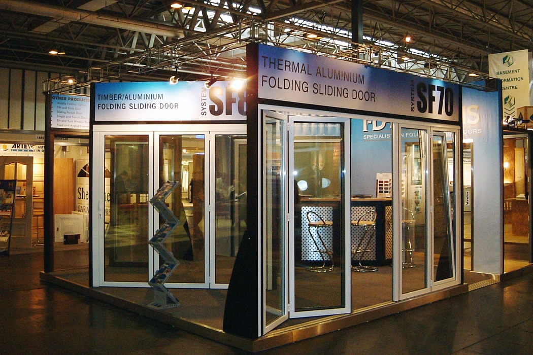 Exhibition booth displaying thermal aluminum and timber aluminum folding sliding doors, with open door panels and informational signs.