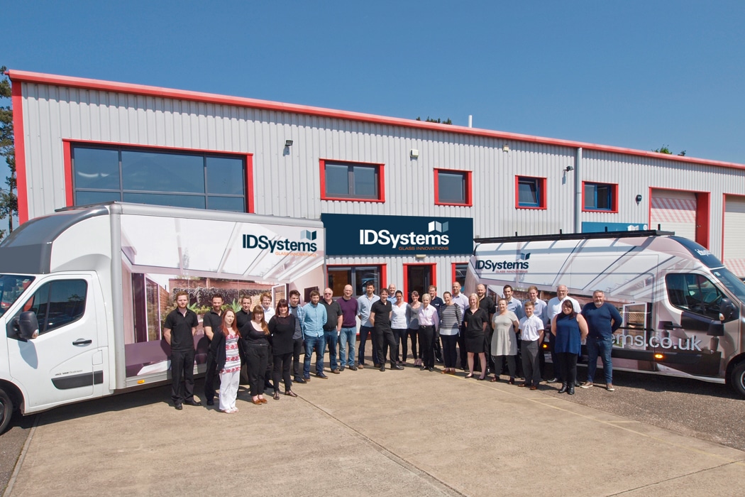 A group of people stands in front of an industrial building with "IDSystems" signs, flanked by two branded vans on a sunny day.