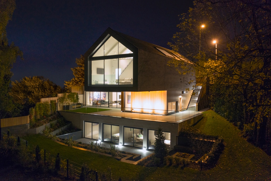 Modern two-story house with large glass windows and sloped roof, illuminated at night, surrounded by landscaped greenery and outdoor lighting.