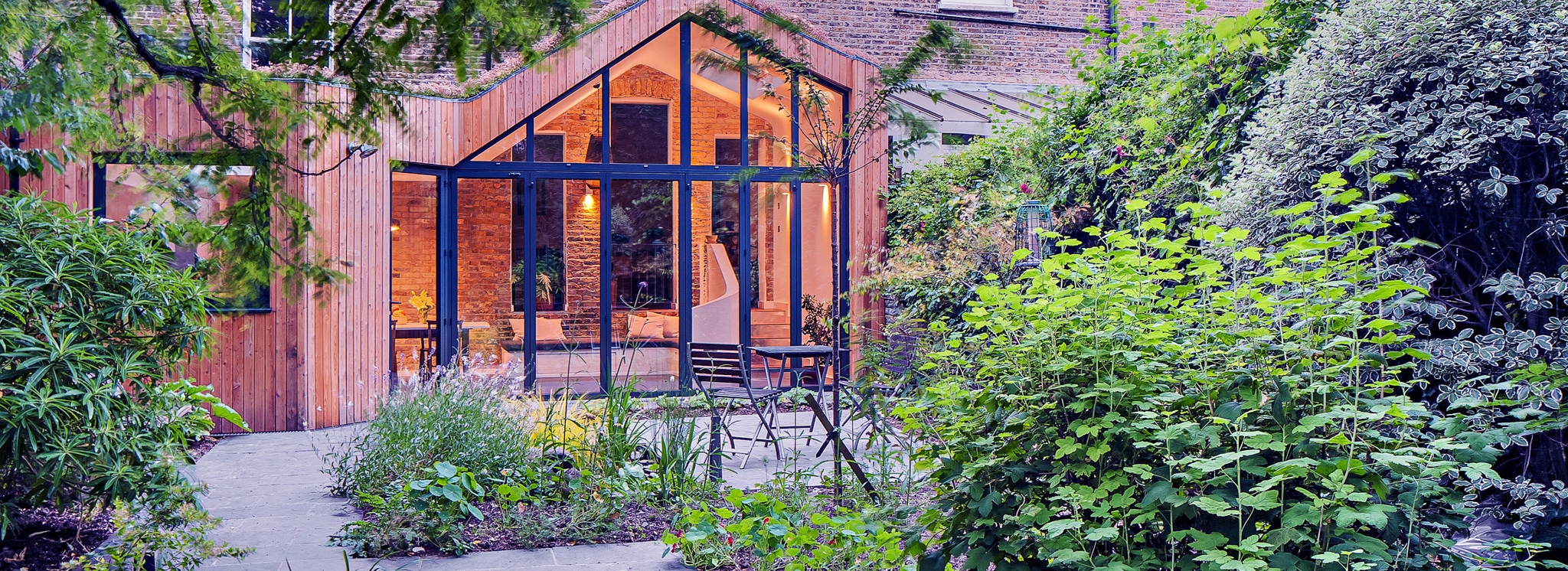 A modern glass-walled extension with wood siding opens onto a lush, green garden with various plants and a stone patio path.