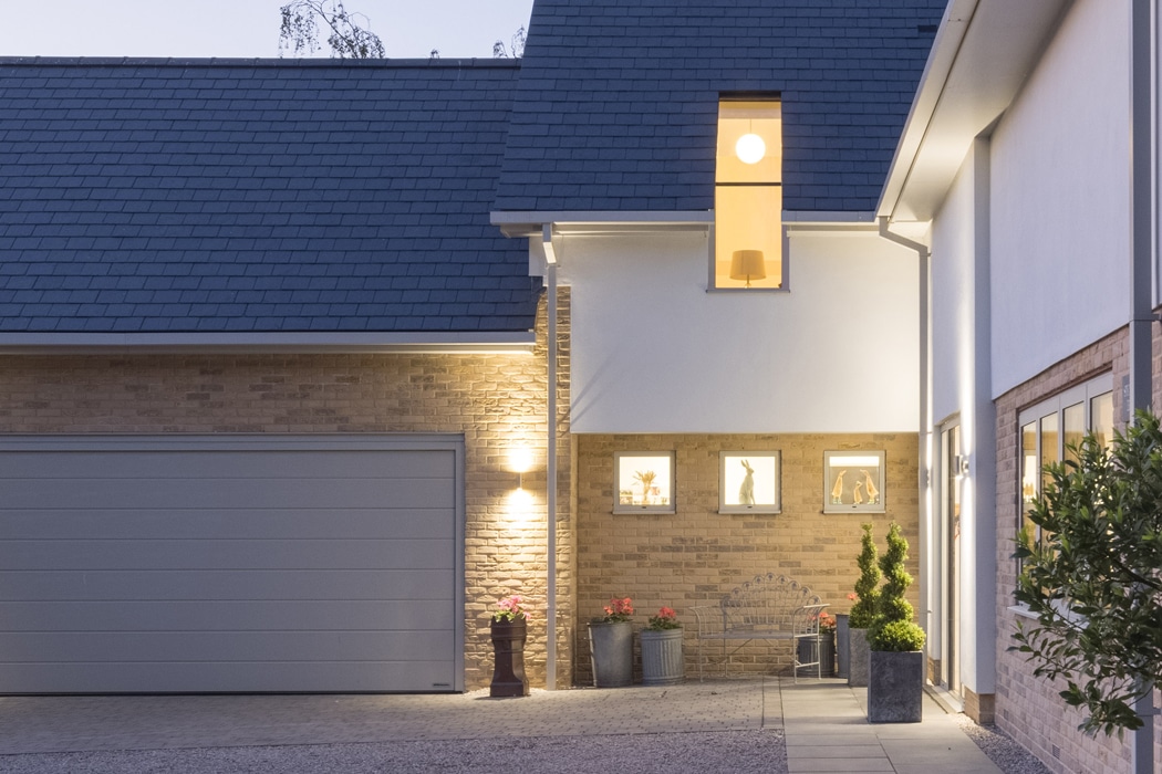 Modern house exterior at dusk with large garage door, illuminated entryway, and three framed pictures on the wall beside potted plants and decorative items.