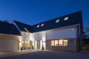 Modern house at dusk with exterior lights on, large windows, brick and white facade, attached garage, and gravel driveway.