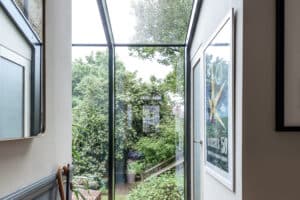 A hallway with glass walls and ceiling offers a clear view of lush trees and a garden outside; framed artwork hangs on the interior walls.