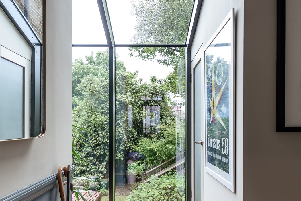 A hallway with glass walls and ceiling offers a clear view of lush trees and a garden outside; framed artwork hangs on the interior walls.