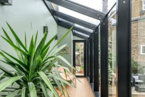 A bright corridor with large glass windows, a green wall, a potted plant, wall lights, a bench, and a door leading outside.