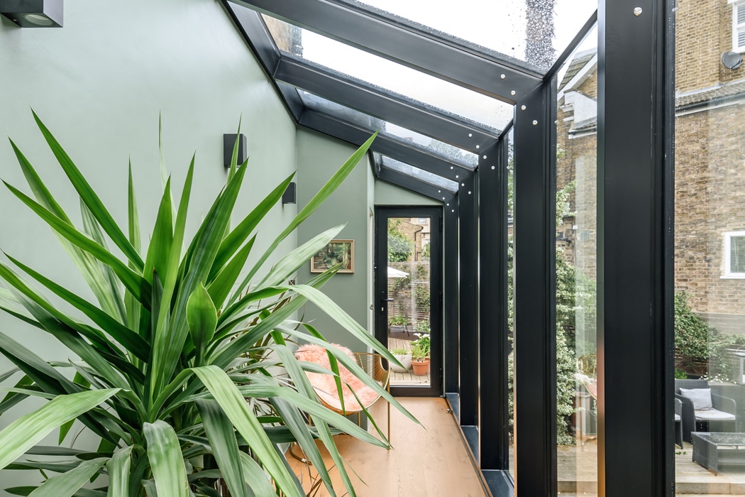 A bright corridor with large glass windows, a green wall, a potted plant, wall lights, a bench, and a door leading outside.