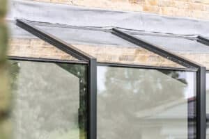 Close-up of a modern glass and metal structure attached to a brick wall, reflecting trees and part of a house in the glass panels.
