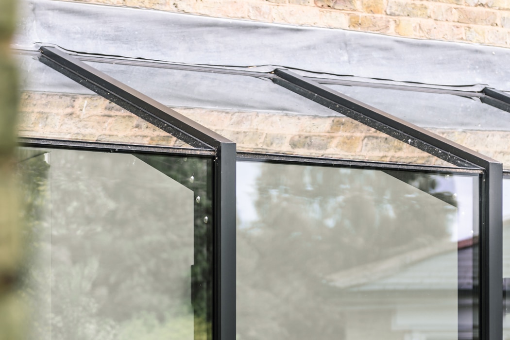 Close-up of a modern glass and metal structure attached to a brick wall, reflecting trees and part of a house in the glass panels.