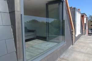 A modern balcony with a frameless glass balustrade and sliding door, showing part of a gray sofa inside and city buildings reflected in the glass.
