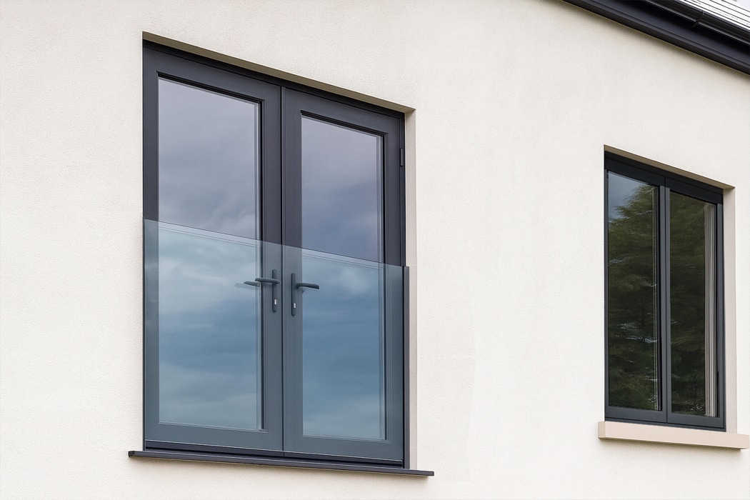 A modern building exterior with a glass-panel French door and a frameless glass balustrade next to a large rectangular window reflecting the sky and trees.