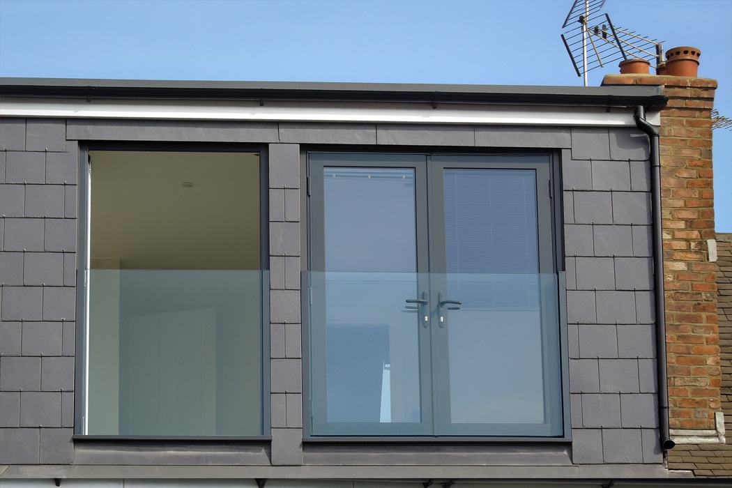 Modern building facade with dark gray tiles, a frameless glass balustrade on the balcony, closed double doors with blue-tinted glass, and a brick chimney with TV antennas on the right side.