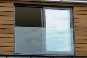 A modern building with wood siding features a large sliding glass window and a sleek frameless glass balustrade, reflecting the cloudy sky.