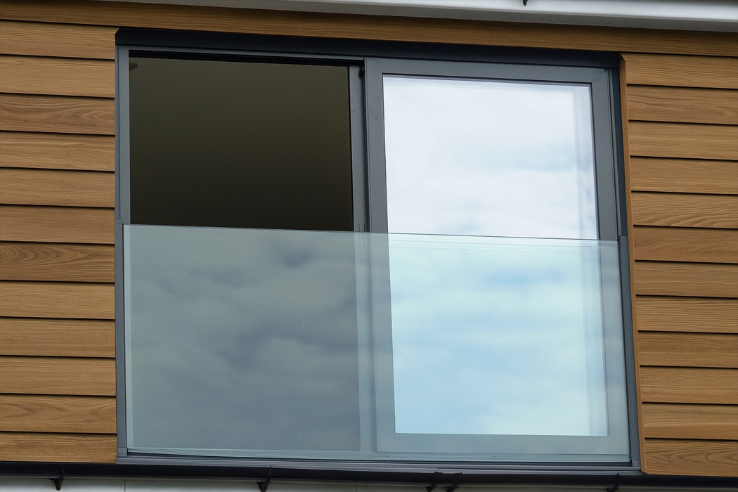 A modern building with wood siding features a large sliding glass window and a sleek frameless glass balustrade, reflecting the cloudy sky.