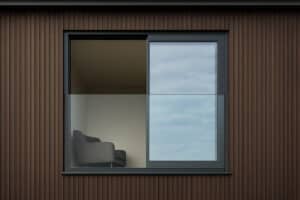 A modern window with a frameless glass balustrade reveals a single gray armchair inside a minimalist room with brown vertical siding on the exterior.