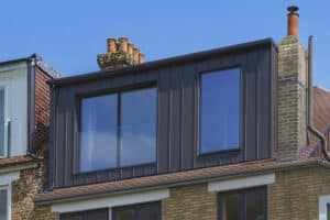 A modern loft extension with large glass windows, a frameless glass balustrade, and dark metal cladding on the upper floor of a brick house, set against a clear blue sky.