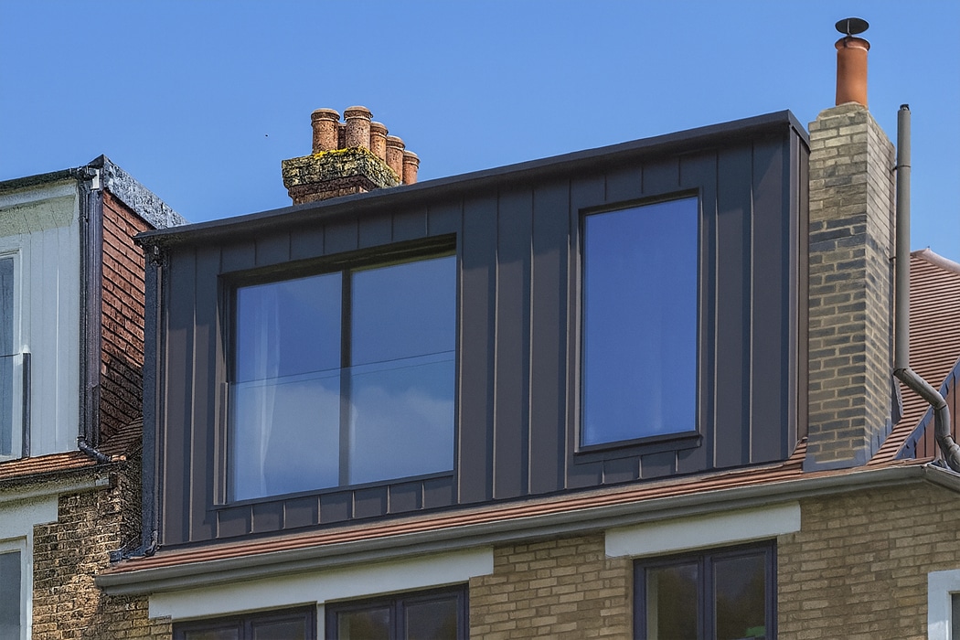 A modern loft extension with large glass windows, a frameless glass balustrade, and dark metal cladding on the upper floor of a brick house, set against a clear blue sky.