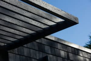 Close-up of a slatted black pergola with raindrops on the surface, attached to a shingled roof under a clear blue sky.