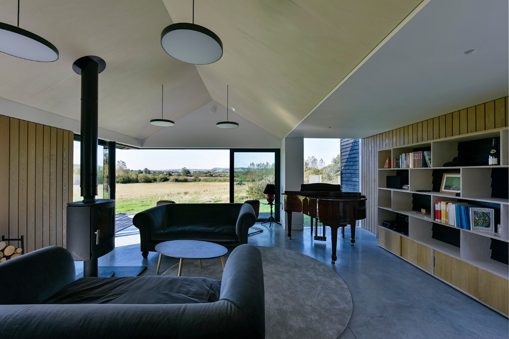 Modern living room with large windows, two dark sofas, a round coffee table, a wood stove, a grand piano, and built-in bookshelves, overlooking a scenic outdoor landscape.