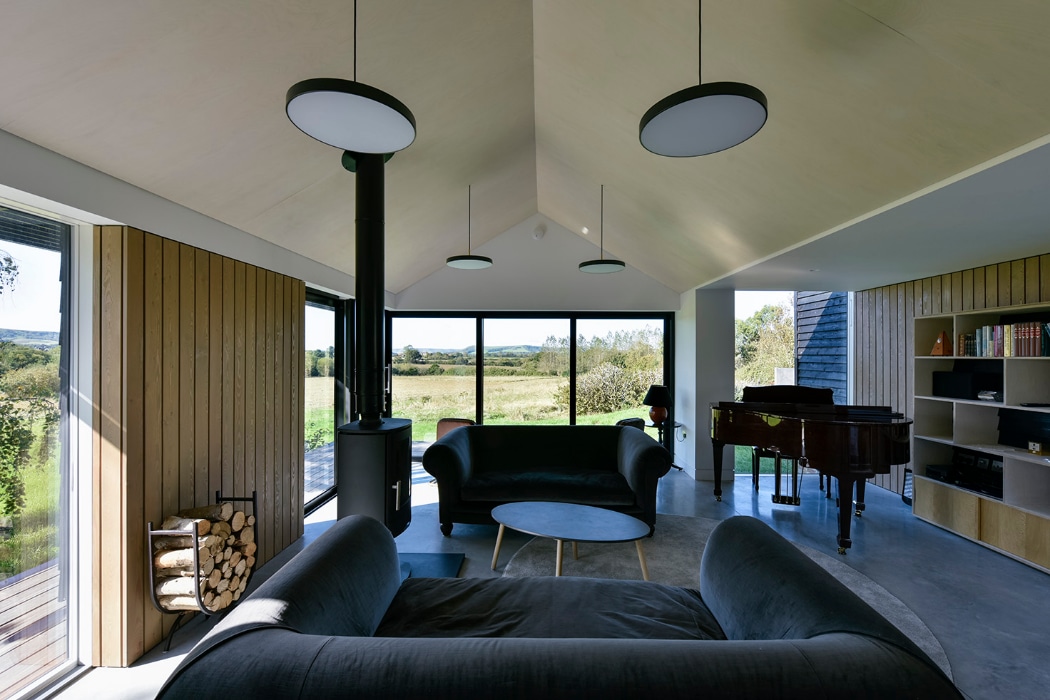 Modern living room with wood paneling, large windows overlooking a scenic landscape, dark sofas, a black wood stove, and a grand piano next to built-in shelves.