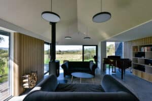Modern living room with wood paneling, large windows, black sofas, a round coffee table, a wood stove, a stack of firewood, bookshelves, and a grand piano overlooking a scenic landscape.