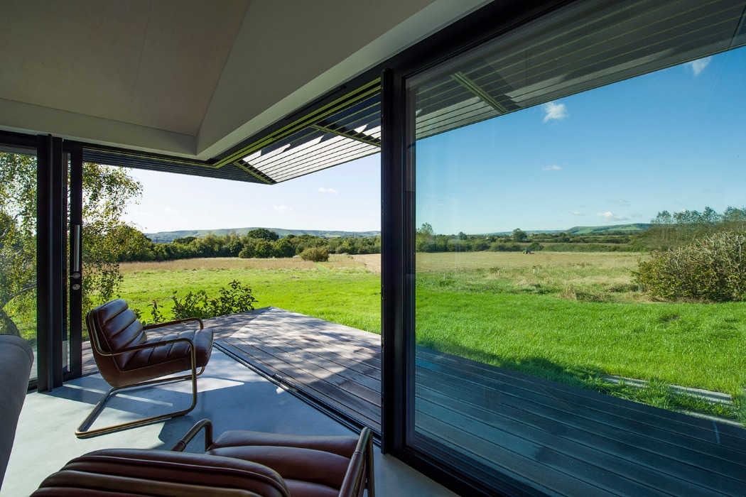 A modern room with large glass walls opens to a patio with two leather chairs, overlooking a grassy field and distant hills under a clear blue sky.
