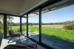 Modern room with large floor-to-ceiling glass windows overlooking a grassy field and distant hills under a clear blue sky. Two brown leather chairs face the view.