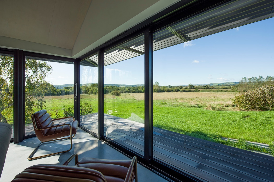 Modern room with large floor-to-ceiling glass windows overlooking a grassy field and distant hills under a clear blue sky. Two brown leather chairs face the view.