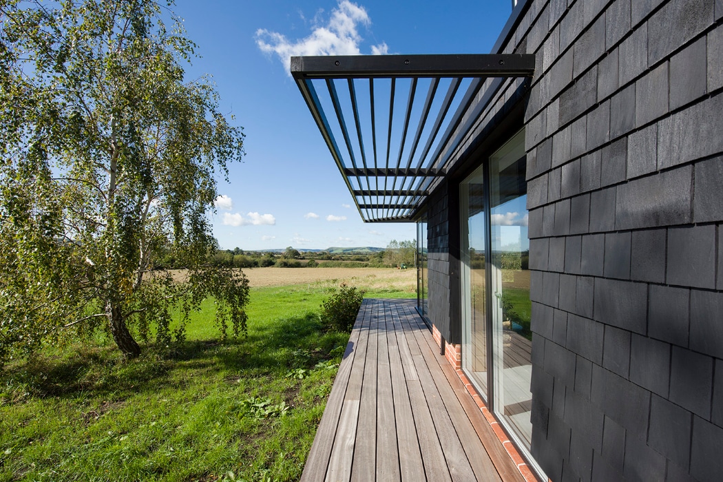 A modern house with dark shingles, a wooden deck, and a metal pergola overlooks a grassy field and distant hills under a blue sky with scattered clouds.