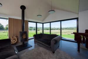 Modern living room with large windows, a wood-burning stove, gray sofas, a round rug, and a view of green fields and hills outside.