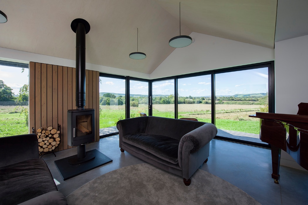 Modern living room with large windows, a wood-burning stove, gray sofas, a round rug, and a view of green fields and hills outside.
