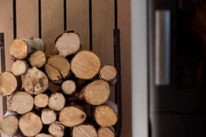 A stack of cut firewood is neatly arranged in a metal log holder next to a wall and a refrigerator.