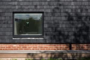A rectangular window set in a dark shingle-clad wall above a brick foundation, with reflections of trees and landscape visible in the glass.