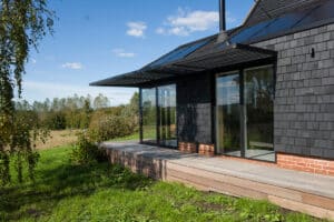 Modern house with dark shingle siding, large glass doors, and a wooden deck, set in a grassy landscape with trees and blue sky in the background.