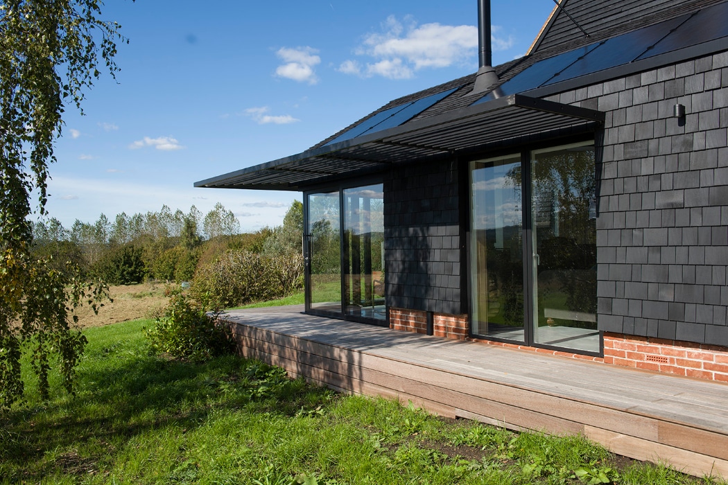 Modern house with dark shingle siding, large glass doors, and a wooden deck, set in a grassy landscape with trees and blue sky in the background.