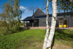 A modern house with dark shingles, large windows, and solar panels sits in a grassy area with birch trees and clear blue sky.