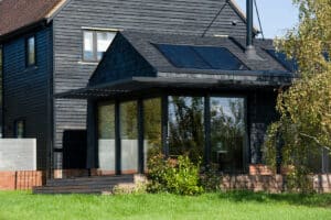 A modern black wooden house with large glass sliding doors, solar panels on the roof, and a small garden in front, set against a grassy lawn.