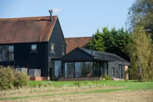 A black wooden house with large windows and a sloped roof sits next to a grassy field, surrounded by trees under a clear blue sky.