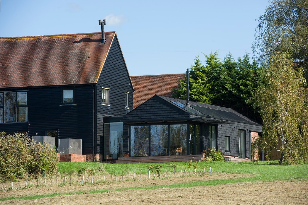 A black wooden house with large windows and a sloped roof sits next to a grassy field, surrounded by trees under a clear blue sky.