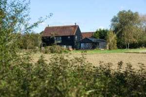 A two-story black house with a sloped roof stands in a rural area, surrounded by trees and an open field under a blue sky.